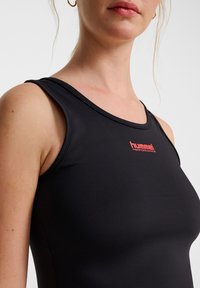 Svart ärmlös träningslinne gjort av slät tyg, med en röd "hummel"-logotyp på bröstet och en figurnära design med rund halsringning.