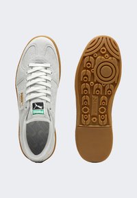 Białe zamszowe sneakersy z siateczkowym językiem, z zielono-czarną metką oraz gumową podeszwą z okrągłymi i liniowymi wzorami.