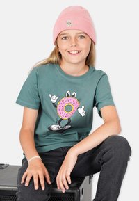 Vihreä lyhythihainen t-paita, jossa on sarjakuvahahmo, joka pitää pinkkiä donitsia. Lapsella on päässään pinkki pipo ja mustat farkut.