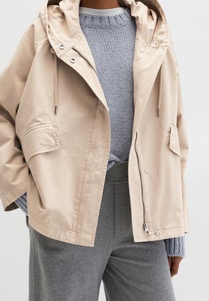Persoon die een beige capuchonjas met voorzakken draagt, een lichtblauwe gebreide trui en grijze broek, getoond van de schouders tot halverwege het dijbeen.