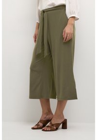 Kvinna i olivgröna vida cropped byxor med tygbälte och bruna klacksandaler, stående mot en enfärgad vit bakgrund.