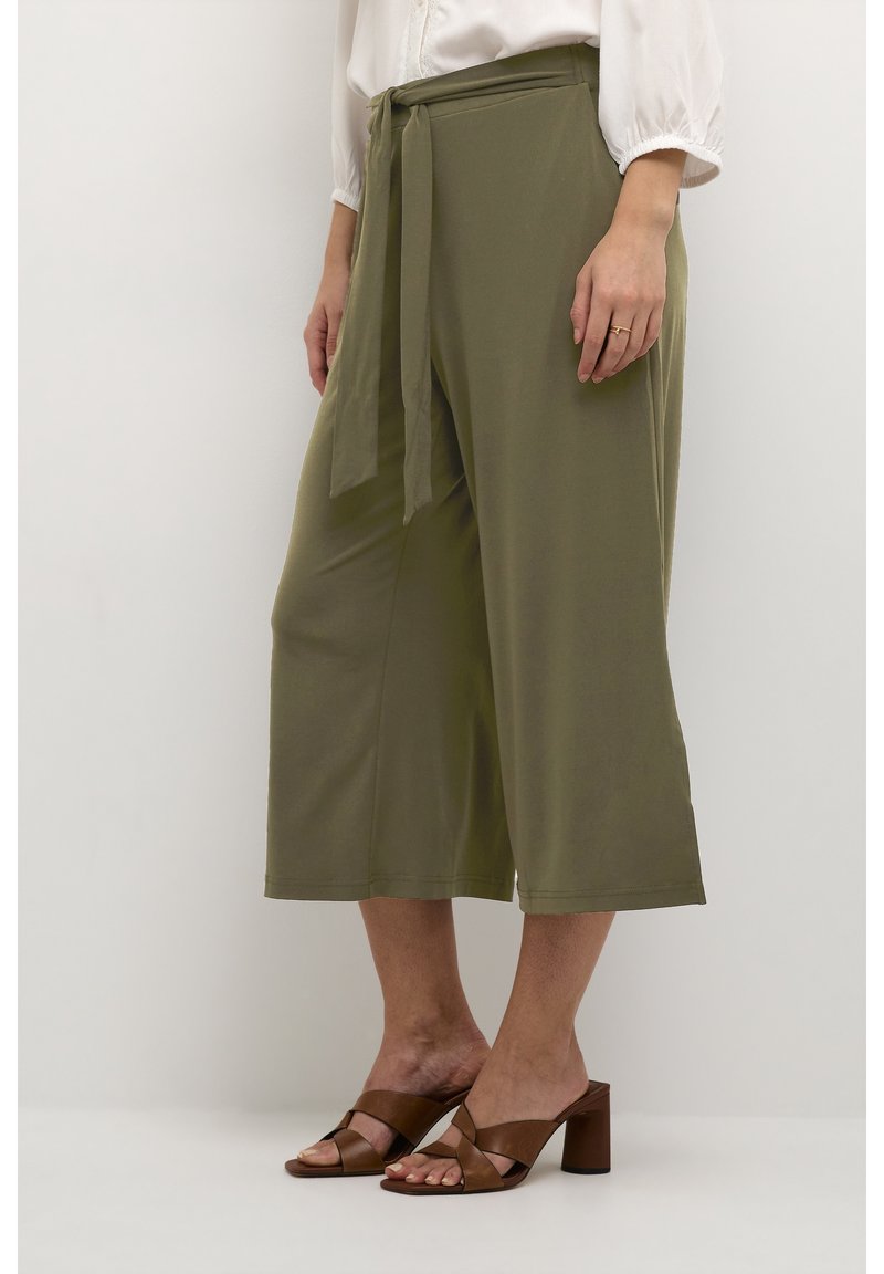 Kvinna i olivgröna vida cropped byxor med tygbälte och bruna klacksandaler, stående mot en enfärgad vit bakgrund.