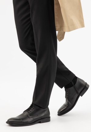 Schwarze Lederschuhe, getragen mit schwarzen Hosen und einem beigen Trenchcoat, Beine übereinandergeschlagen, schreitend vorwärts auf weißem Hintergrund.