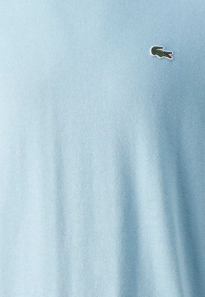 Pull en coton bleu clair avec une texture lisse, arborant un petit logo vert et rouge sur la poitrine.