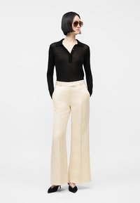 Kvinne med kort svart hår iført store solbriller, en gjennomsiktig svart langermet topp, kremfargede høytlivs vide bukser og svarte hæler.