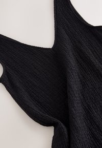 Haut noir sans manches en tissu texturé avec un motif ondulé, présenté à plat sur une surface claire.