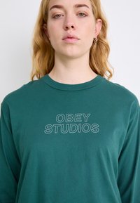 Zeleni dukserica dugih rukava s natpisom "OBEY STUDIOS" u bijelom na prednjoj strani. Napravljen od mekog pamuka, ima klasičan okrugli izrez.