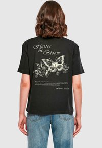Sort bomulds T-shirt med et stort hvidt sommerfuglegrafik og blomstrende accenter på ryggen, med tekstoverlay, der beskriver naturtemaer.
