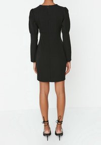 Svart figursydd klänning med långa ärmar, puffiga axlar och dragkedja i ryggen. Slät textur, knälång design, som kombineras med svarta strap-upp klackar.