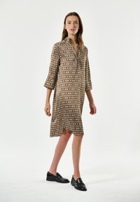 Braun gemustertes Hemdkleid mit Kragen, Dreiviertelärmeln und seitlichen Schlitzen. Kombiniert mit schwarzen Loafers. Glatte Stoffstruktur.