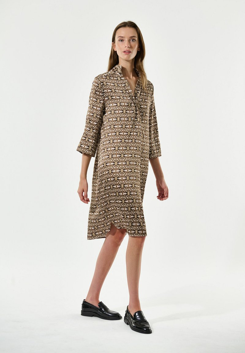 Vestito camicia marrone a fantasia con colletto, maniche a tre quarti e spacco laterale. Abbinato a mocassini neri. Tessuto dalla superficie liscia.