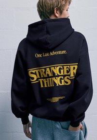 Černá mikina s kapucí, na zadní straně s nápisem "STRANGER THINGS" žlutým písmem a frází "Jedno poslední dobrodružství." Měkčená látka, oversized střih.