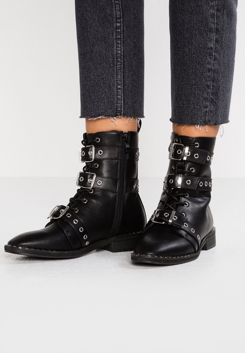 Bottines en cuir noir avec trois boucles et œillets argentés, portées avec un jean noir raccourci et effiloché, sur un sol blanc.