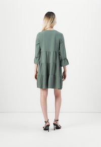 Robe verte à volants avec des manches trois-quarts, des poignets à volants et une coupe ample. Portée avec des talons hauts noirs à lanières ornés de pois.