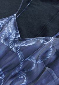 Gros plan d'une robe bleue à fines bretelles avec un motif de corde blanche, posée sur un fond de tissu foncé.