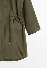 Olivgrüne Langarmjacke mit tailliertem Schnitt, Klappentaschen, Kordelzug und verstellbaren Manschetten vor weißem Hintergrund dargestellt.