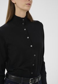 Fekete kötött kardigán magas gallérral, elöl gombokkal és hét fém gombbal. Sötét farmernadrág fölött viselve, fekete övvel.