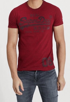 Homme portant un t-shirt rouge moulant Superdry avec des textes et des chiffres noirs, associé à un jean bleu foncé.