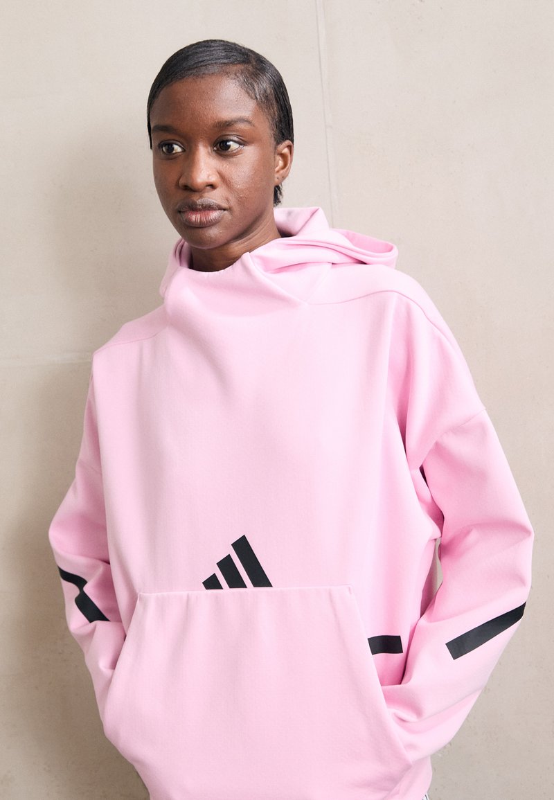 Rožinė kapučinė striukė, pagaminta iš minkštos medžiagos, su dideliu priekiu kišeniu, juodomis juostelėmis ant rankovių ir Adidas logotipu priekyje.