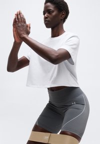 Άτομο που φοράει άσπρο κοντό μπλουζάκι και γκρι σορτς γυμνάζεται με μπεζ λαστιχένια ταινία αντίστασης γύρω από τους μηρούς, τα χέρια πλεγμένα μπροστά.