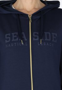 Navy zip-up hoodie made from textured fabric. Features a gold zipper and printed "SEA SIDE NAUTICAL LEGACY" in dark lettering on chest.