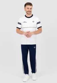 Balta T-kreklā ar tumši ziliem svītrām un logo, apvienota ar tumši ziliem biksēm ar baltām sānu svītrām. Modelis valkā baltas sporta kurpes.