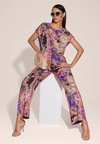 Multicolore jumpsuit met een paisleypatroon in paars, zwart en goud, gecombineerd met doorzichtige sandalen met hakken en een clutch.