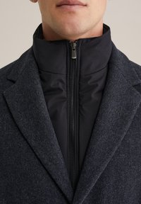 Cappotto di lana navy con colletto alto. Lo strato interno presenta un tessuto imbottito nero con una zip centrale e hardware argentato. Finitura testurizzata.