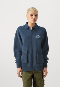 Námornícky modrá mikina na zips vyrobená z mäkkého materiálu, s dvomi prednými vreckami a vyšitým textom "MARATÓN ŽIVOTA" na hrudi.