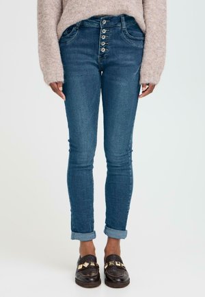 Schmale Jeans aus dunkelblauem Denim mit einer Fünf-Knopf-Front, mit umgeschlagenem Saum und Taschen. Kombiniert mit einem hellen Strickpullover und Loafers.