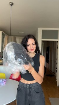 Schwarzes Kleidungsstück in einer durchsichtigen Plastiktüte, mit glatter Textur. Model trägt ein enges schwarzes Oberteil und hoch taillierte graue Jeans.