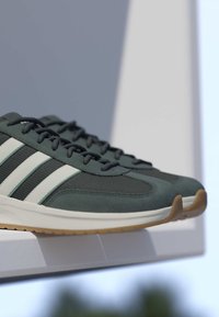 Chaussures de sport vert foncé avec des bandes blanches et des semelles en gomme présentées sur une surface blanche inclinée sur un fond ombragé.