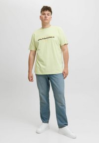 Hellgrünes T-Shirt mit kurzen Ärmeln und schwarzem sowie goldenem "JACK&JONES"-Logo, kombiniert mit hellblauen Jeans und weißen Sneakers. Einfaches Design.