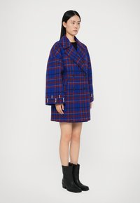 Casaco de lã azul e vermelho em padrão xadrez, com um design de botão duplo, gola larga e mangas oversized. Combinado com botins pretos até ao tornozelo.