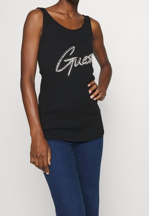 Personne portant un haut noir sans manches avec le logo en strass "Guess" et un jean bleu, debout avec une main sur la hanche.