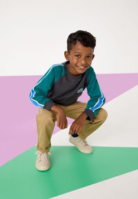 Graues Langarmshirt mit türkisfarbenen Akzenten und blauen Streifen, khakifarbene Hose und helle Sneaker; Junge hockt auf farbenfrohem, geometrischem Hintergrund.