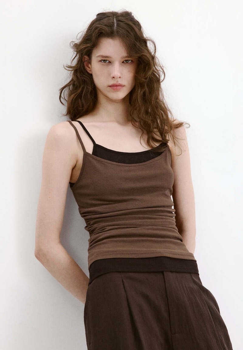 Jeune femme aux cheveux bruns ondulés portant plusieurs débardeurs marron superposés et un pantalon marron ample, debout contre un mur blanc.