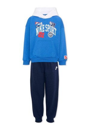 Niebieska bluza Nike z białym kapturem i różnymi logo, w zestawie z ciemnoniebieskimi spodniami dresowymi Nike z białym logo na lewej nodze.