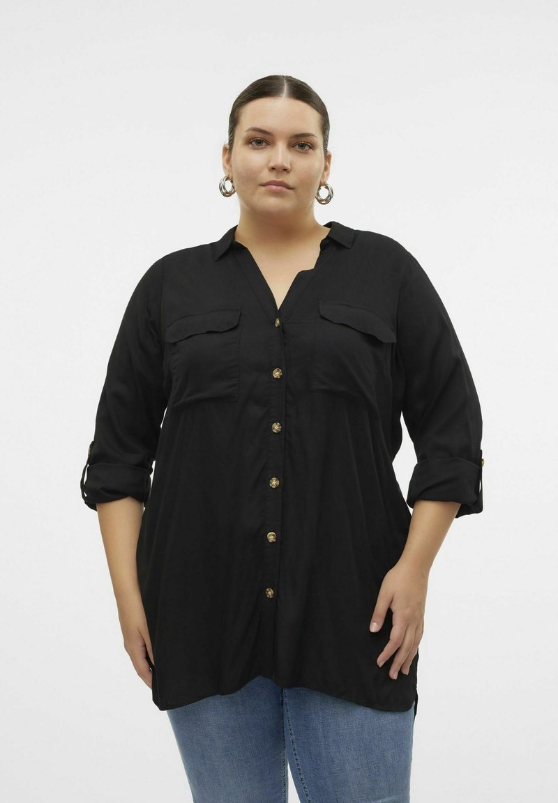 Sort skjorte med knapper og krave, to brystlommer, guldknapper, opdragne ærmer og en løs pasform, parret med lyseblå jeans.