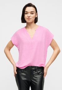 Νεαρή γυναίκα με κοντά καστανά μαλλιά φοράει ροζ πλεκτό τοπ με V ντεκολτέ και μαύρο δερμάτινο παντελόνι, στέκεται με τα χέρια στη μέση.