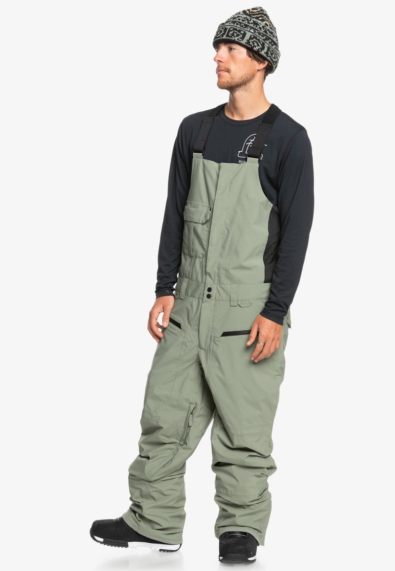 Salopette de ski en tissu vert clair avec des bretelles ajustables, des poches avant et des accents zippés. Associée à un bonnet en tricot à motifs.