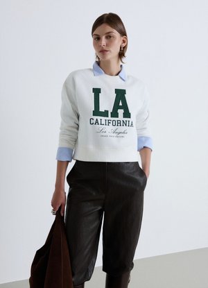 Donna che indossa una felpa bianca con testo "LA California", camicia azzurra con colletto, pantaloni neri e che tiene una borsa marrone scuro.