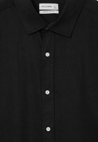 Schwarze Hemdbluse mit weißen Knöpfen, spitzem Kragen und einem Etikett, das die Marke "Calliope", die Größe "L" und "Reguläre Passform" zeigt.