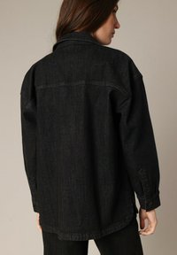 Zwarte spijkerjack met een losse pasvorm, voorzien van een puntige kraag, knoopmanchetten en een gebogen zoom. Zachte textuur en minimale details.