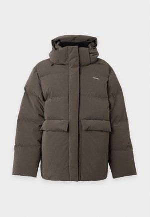 Femme vêtue d'une doudoune vert foncé, d'un pull à col roulé beige, d'un jean évasé bleu et de chaussures foncées, debout sur un fond blanc.