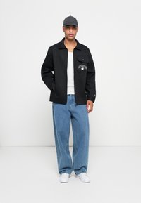Svart jakke med logo-patch og enkelt brystlomme, kombinert med lyseblå vide jeans og hvite sneakers. Grå caps.
