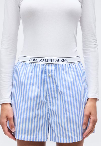 Personne portant un boxer à rayures verticales bleues et blanches avec une ceinture blanche portant l'inscription "Polo Ralph Lauren" et une chemise blanche à manches longues.