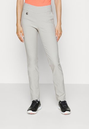 Persoon draagt lichtgrijze slim-fit broek, oranje top en zwarte sportschoenen, staand tegen een effen witte achtergrond.