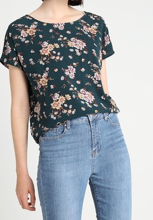 Vrouw draagt een donkergroene bloemendessin korte mouw top gecombineerd met lichtblauwe high-waisted jeans tegen een effen achtergrond.