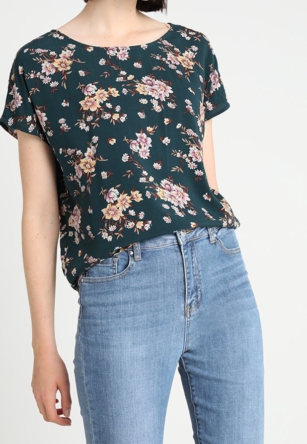 Femme portant un haut court à manches courtes fleuri vert foncé associé à un jean taille haute bleu clair sur un fond uni.
